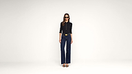 Woman wearing sunglasses, a black long-sleeve top, dark blue wide-leg jeans, a belt, and black heels stands against a plain white background.
