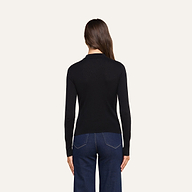 A woman with long brown hair, dressed in a slim fit black Sterling Sweater and dark blue jeans, stands facing away from the camera against a light background.