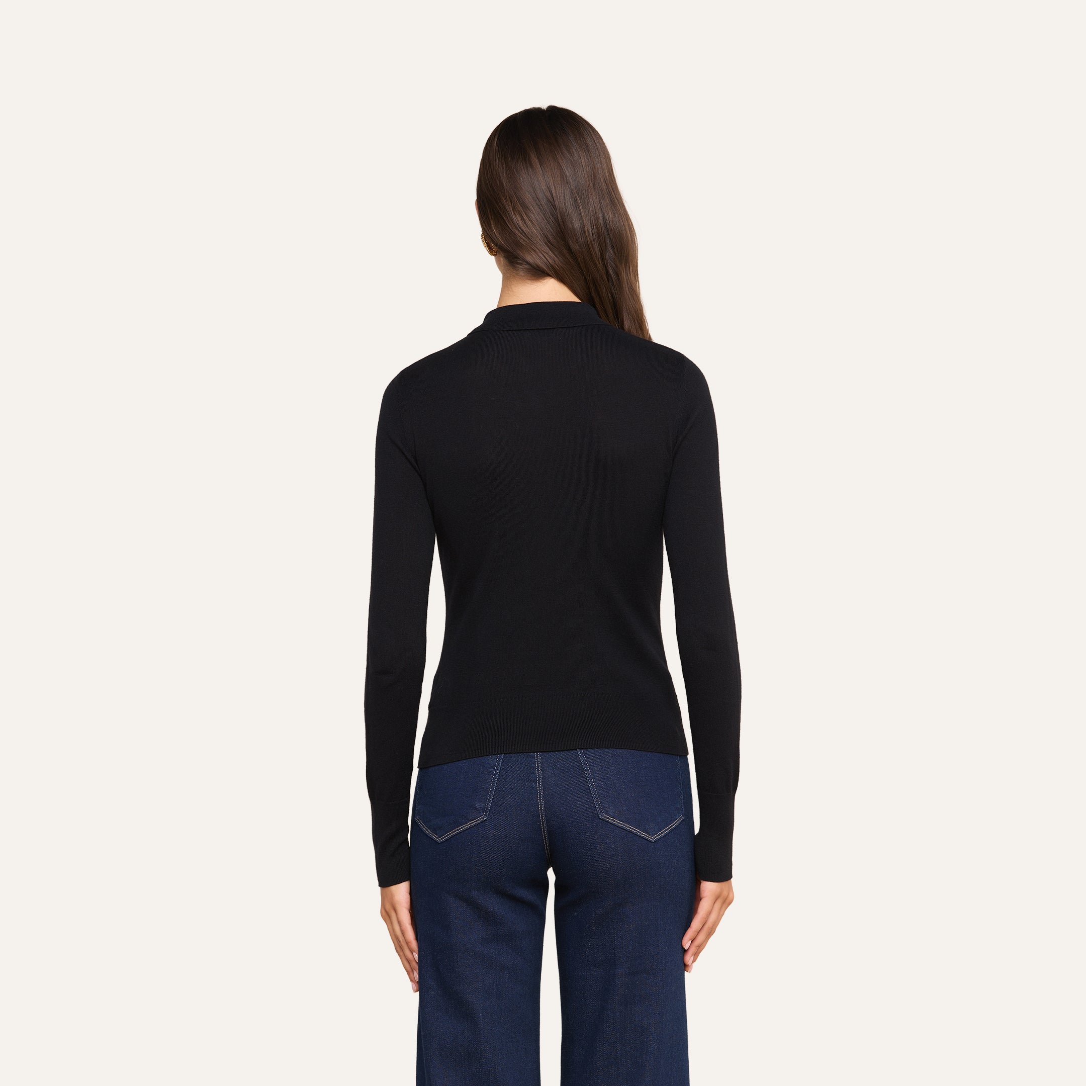 A woman with long brown hair, dressed in a slim fit black Sterling Sweater and dark blue jeans, stands facing away from the camera against a light background.