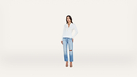 A woman stands against a plain white background, wearing a white long-sleeve top, light blue ripped jeans, and heels.
