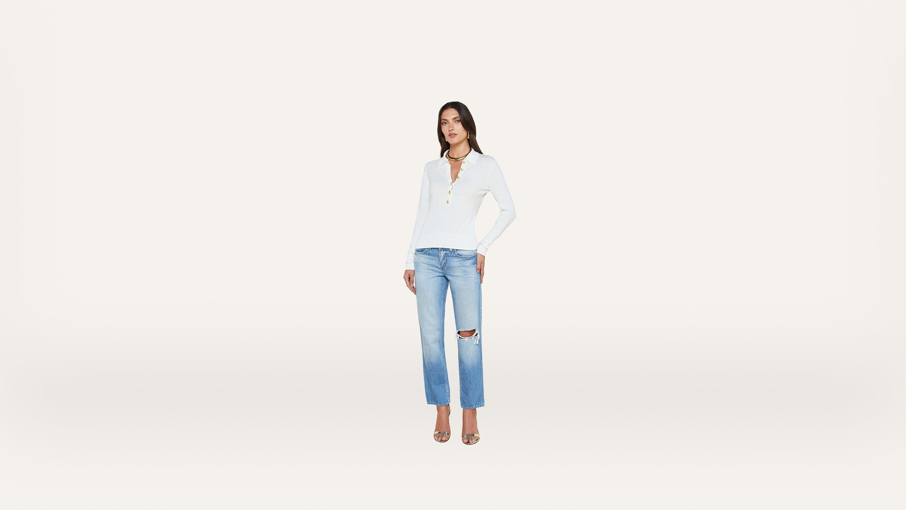 A woman stands against a plain white background, wearing a white long-sleeve top, light blue ripped jeans, and heels.