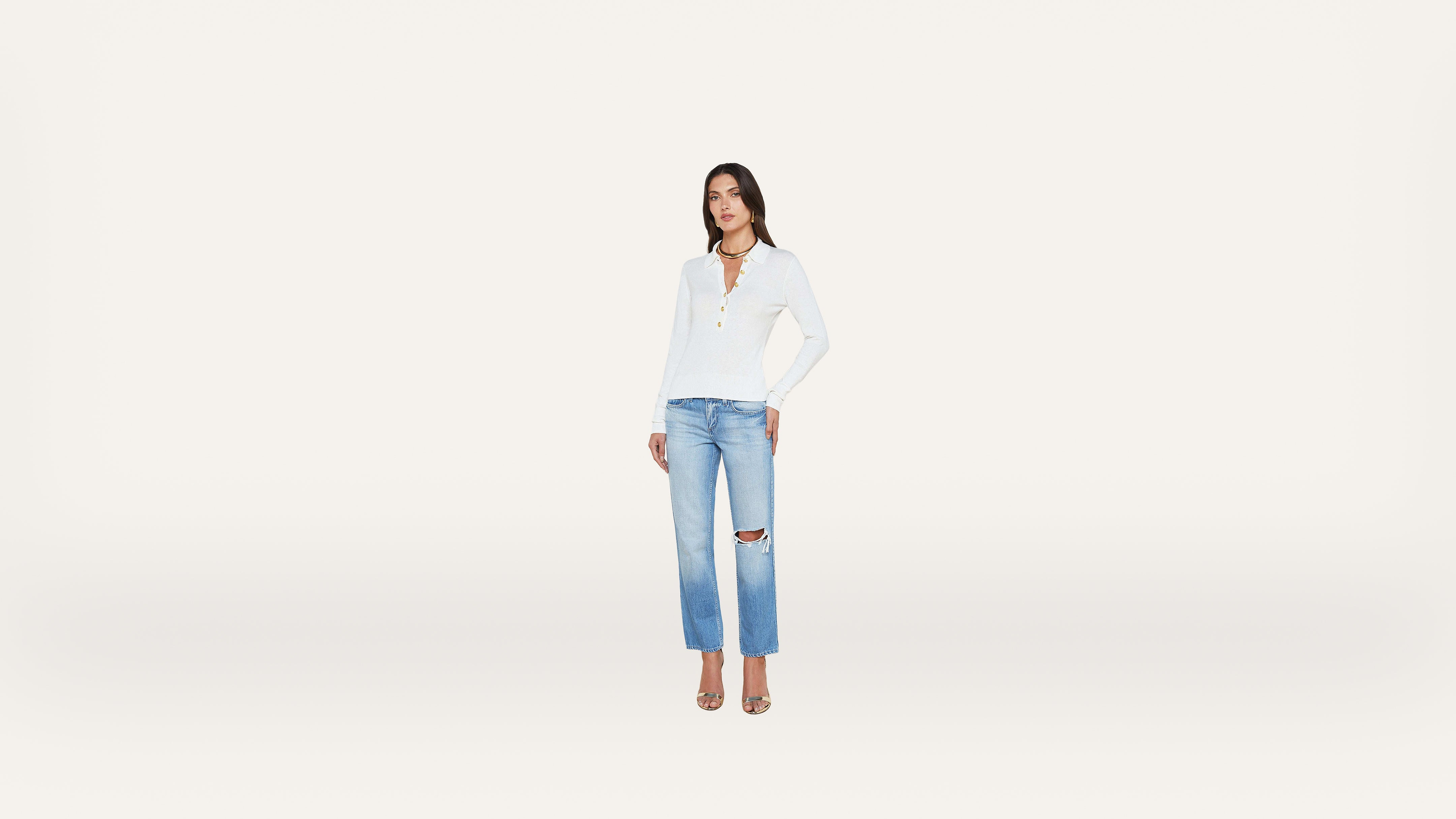 A woman stands against a plain white background, wearing a white long-sleeve top, light blue ripped jeans, and heels.