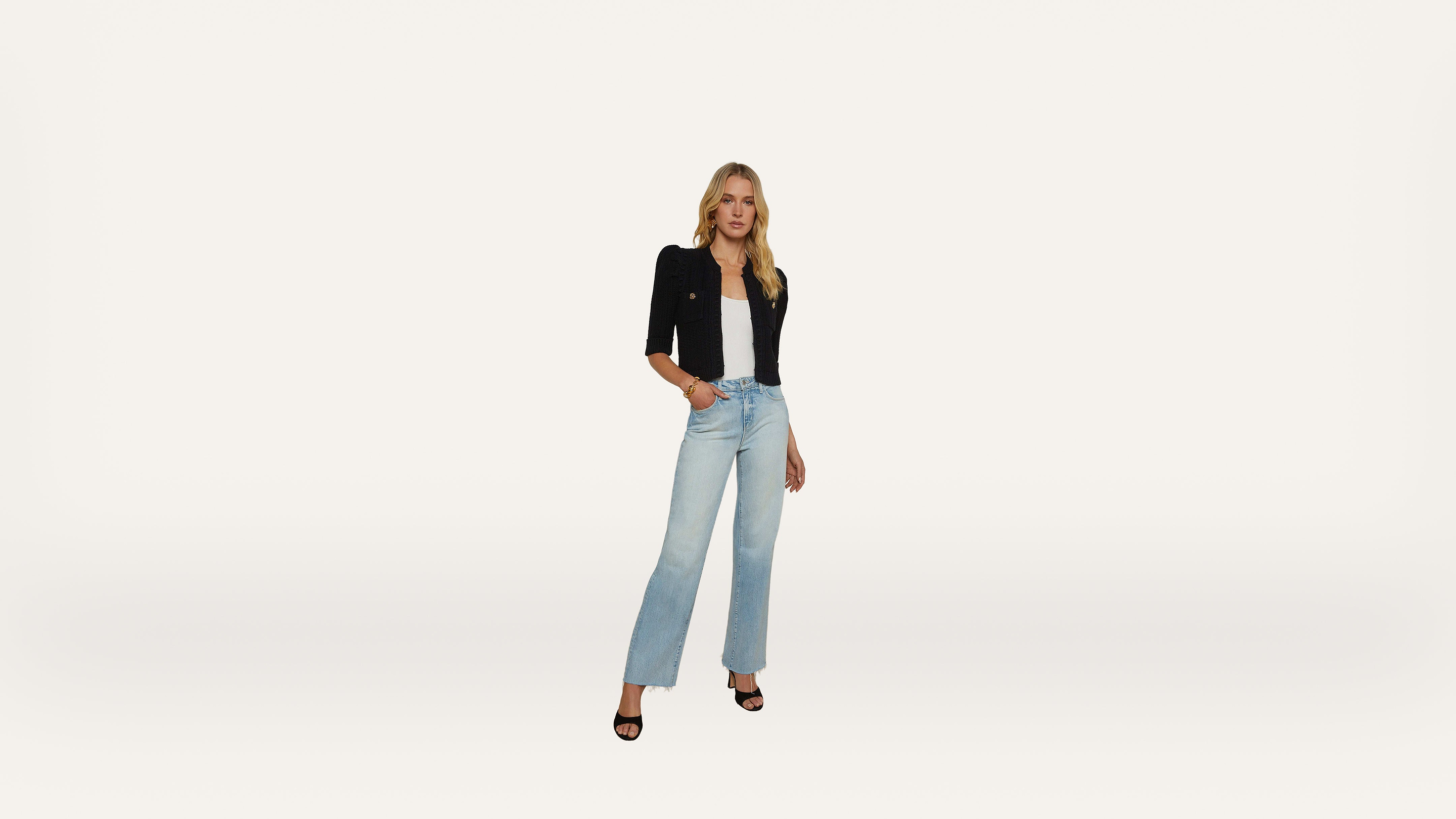 A woman stands against a plain white background, wearing a black jacket, white top, light blue high-waisted jeans, and black open-toe heels.