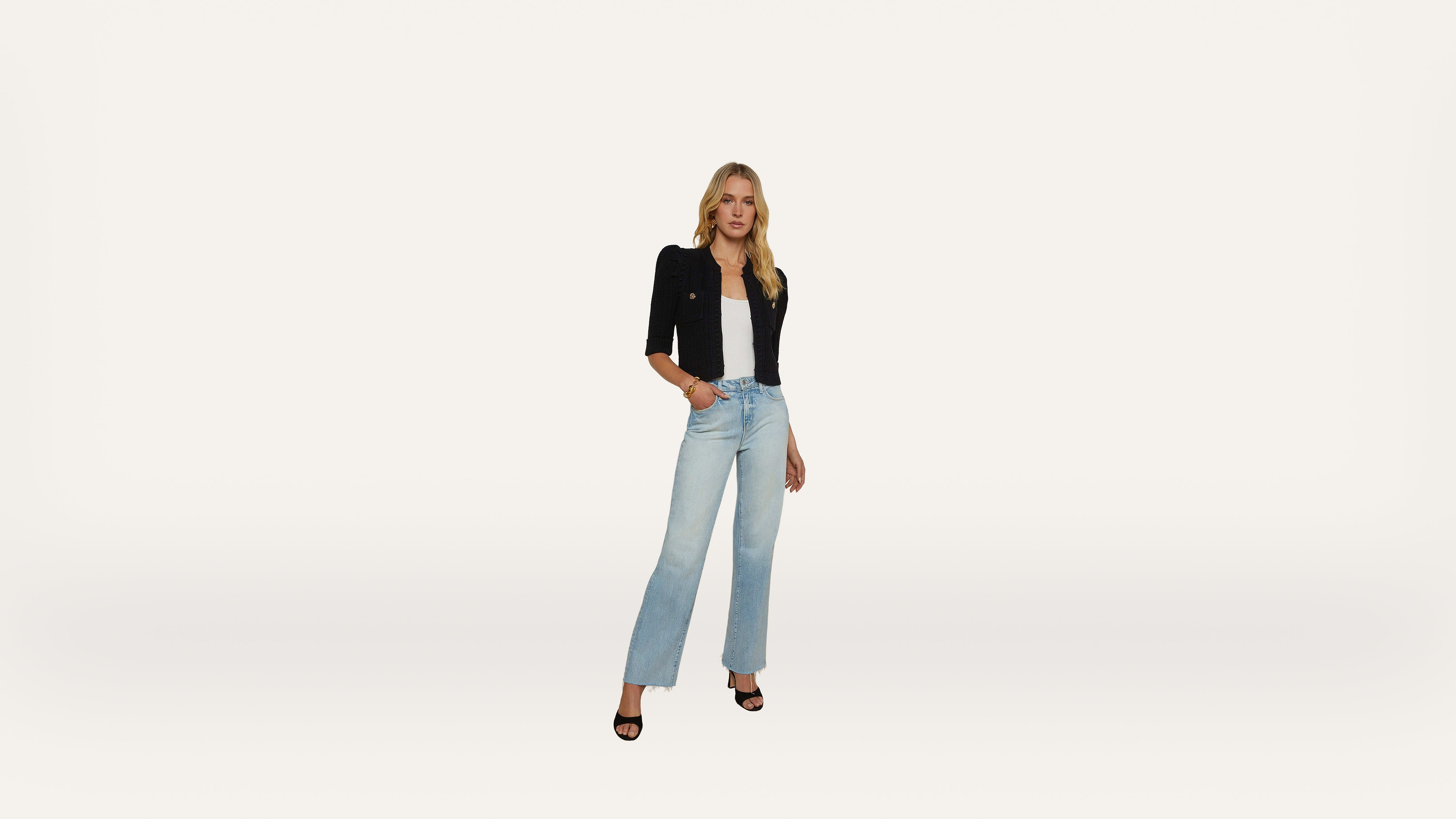 A woman stands against a plain white background, wearing a black jacket, white top, light blue high-waisted jeans, and black open-toe heels.