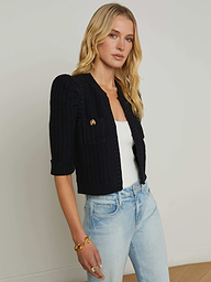 A woman with long blonde hair wears the Delilah Cropped Cardigan, a black textured knit, layered over a white top and paired with light blue jeans as she poses against a plain light background.