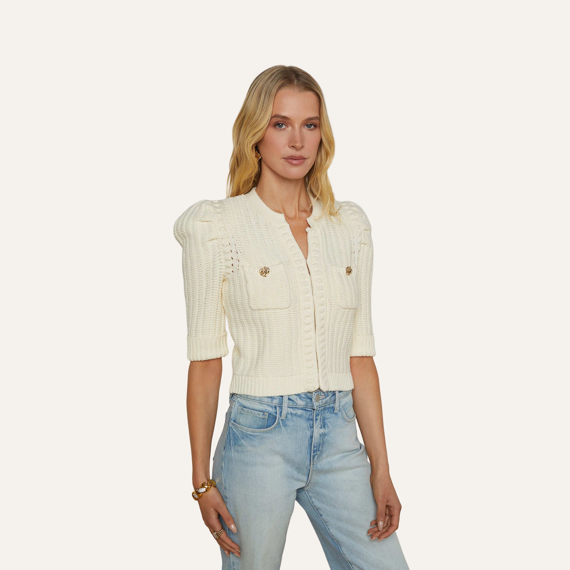 A woman with blonde hair wears the Delilah Cropped Cardigan, a vintage white textured knit style with front pockets, paired with light blue jeans. She stands against a plain, light-colored background and has a neutral expression.
