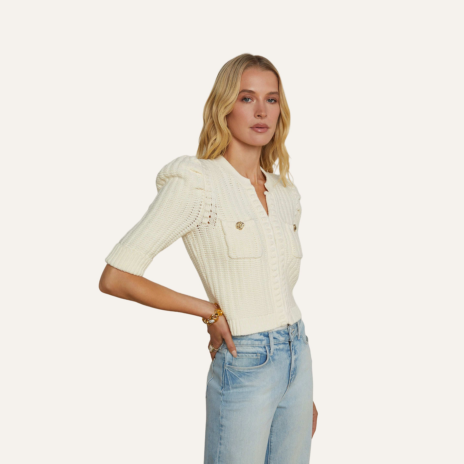 A woman with blonde hair wears the Delilah Cropped Cardigan, a white textured knit with front pockets, paired with light blue jeans. She stands with one hand in her pocket against a plain background.