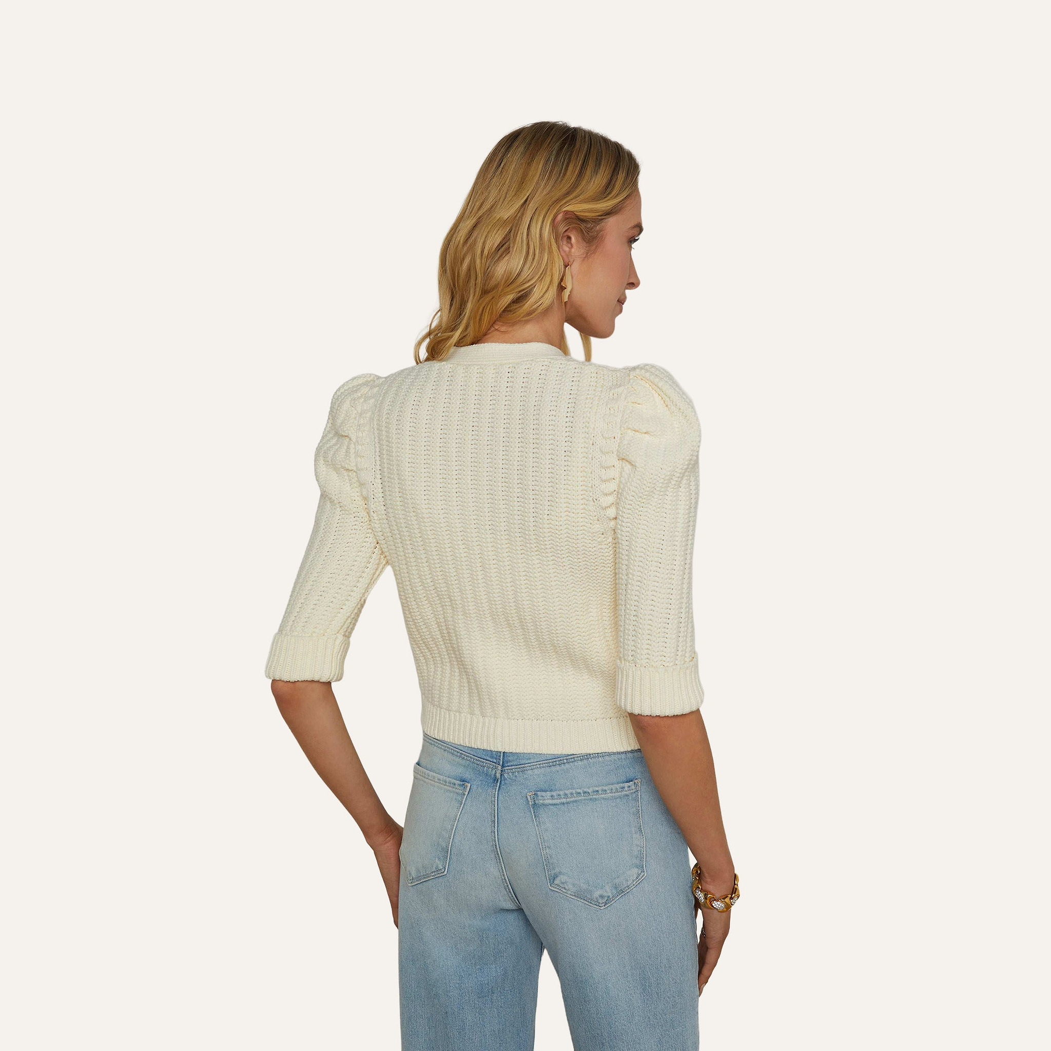 A woman with blonde hair is shown from the back against a plain, off-white background wearing the Delilah Cropped Cardigan, which features textured knit fabric and puff sleeves, paired with light blue jeans.
