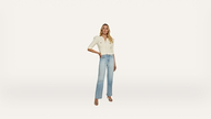 A woman stands against a plain white background, wearing a cream-colored top, light blue high-waisted jeans, and heeled sandals. One hand rests on her hip, and she faces forward, posing confidently.
