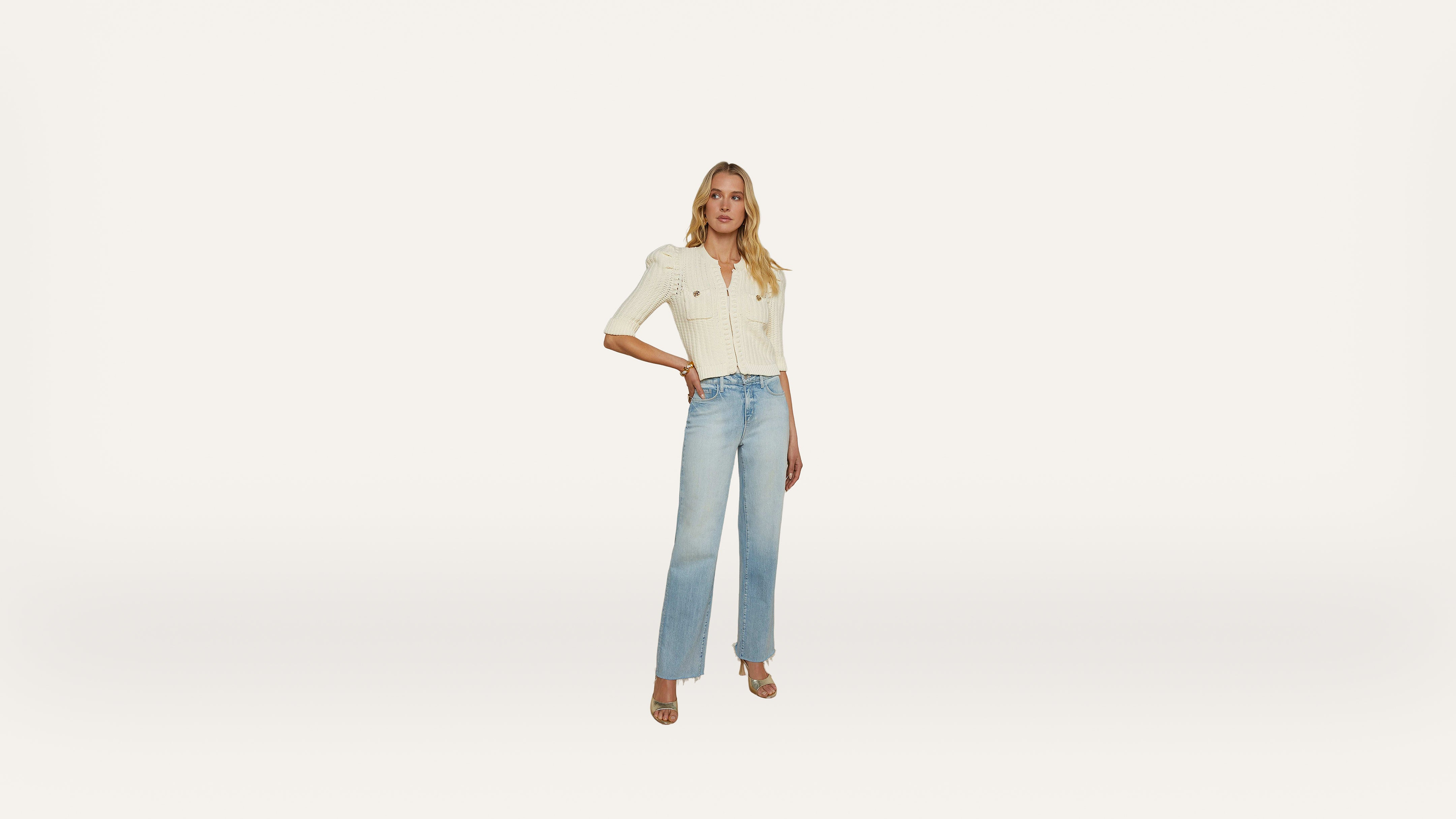 A woman stands against a plain white background, wearing a cream-colored top, light blue high-waisted jeans, and heeled sandals. One hand rests on her hip, and she faces forward, posing confidently.