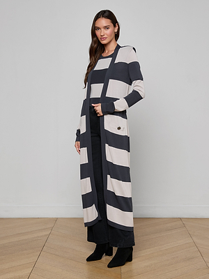 A woman with long dark hair stands indoors against a plain wall, wearing the Berlin Duster Cardigan with graphic stripes over a matching top, black pants, and heeled boots. She appears relaxed.