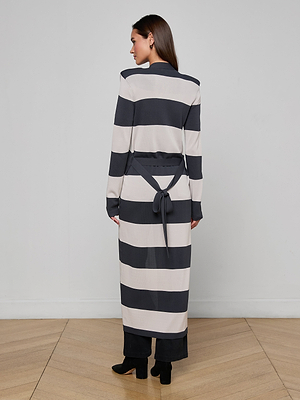 A woman with long brown hair wears the Berlin Duster Cardigan featuring graphic stripes and a belt tied at the back, styled with black pants and heeled boots. She stands on a light wood floor against a plain wall, facing away from the camera.