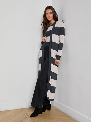 A woman stands against a white wall wearing the Berlin Duster Cardigan with graphic stripes, a matching striped top, black wide-leg pants, and black heeled boots.
