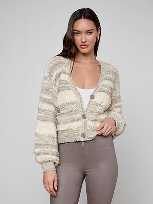 A woman with long dark hair wears the Harriet Cardigan, a cozy wool-blend knit with blouson sleeves, layered over a white top and taupe pants, standing against a plain light background.