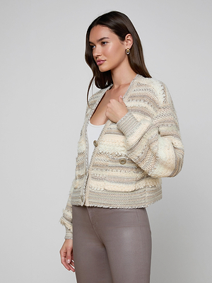 A woman with long brown hair wears the Harriet Cardigan, a cozy wool-blend knit with blouson sleeves, over a white top and taupe pants. She stands sideways, holding the cardigan closed against a plain light background.