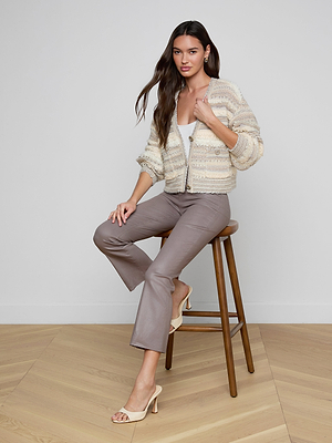 A woman with long brown hair sits on a wooden stool, wearing the Harriet Cardigan with blouson sleeves over a white top, taupe pants, and beige heeled sandals, posing against a plain white wall on a wooden floor.