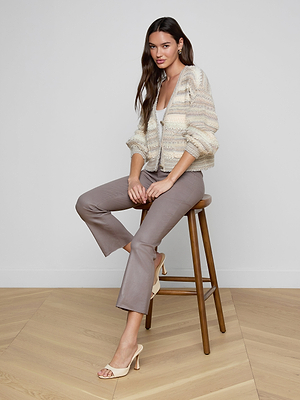 A woman with long brown hair, wearing the Harriet Cardigan with blouson sleeves, taupe cropped pants, and beige heeled sandals, sits on a wooden stool in a minimalist room with light wood floors and white walls.