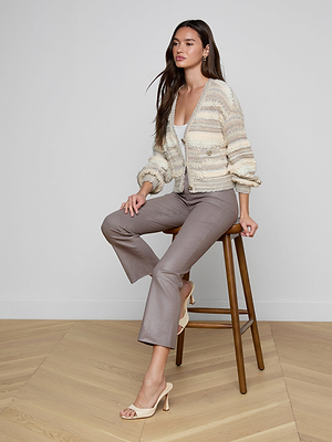 A woman with long brown hair sits on a wooden stool against a plain wall, wearing the Harriet Cardigan with blouson sleeves, a white top, taupe pants, and beige heeled sandals. She poses with one hand on her knee and the other on the stool.