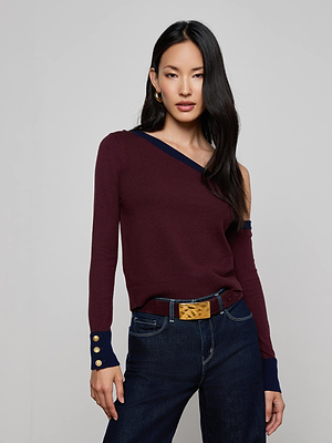 A woman with long black hair models the Maeva One-Shoulder Top by L'AGENCE—burgundy with navy trim—styled with dark slim fit jeans, a brown belt with a gold buckle, and gold hoop earrings. She poses with one hand in her pocket.