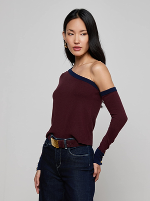 A woman with long dark hair models the L'AGENCE Maeva One-Shoulder Top, a slim-fit maroon style with navy trim, paired with dark jeans. She stands against a plain light background, gazing left with one hand on her hip.