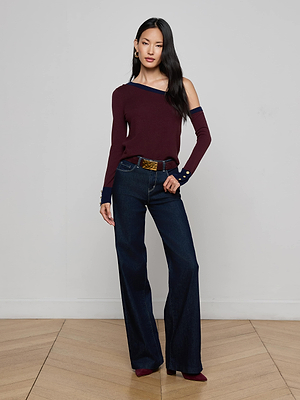 A woman with long black hair wears the L'AGENCE Maeva One-Shoulder Top in burgundy, paired with dark wide-leg jeans and matching shoes, standing indoors on a wooden floor against a plain light-colored wall.