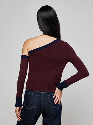 A woman with long dark hair, seen from behind, wears the L'AGENCE Maeva One-Shoulder Top—a slim fit burgundy knit with navy trim—paired with dark jeans and one hand on her hip.