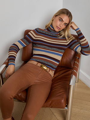 A woman with long blonde hair sits on a brown leather chair, relaxing in the L'AGENCE Olene Turtleneck—featuring ribbed knit in blue, brown, and beige—and high-waisted belted brown pants.