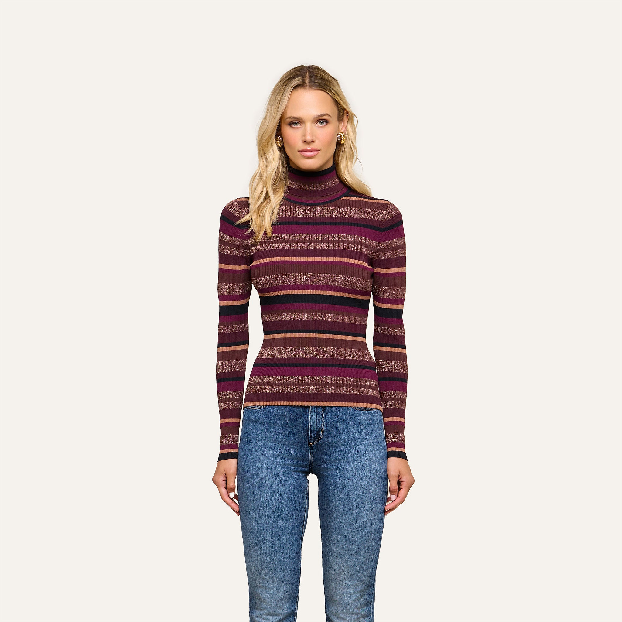 A woman stands against a plain background, wearing the Olene Striped Turtleneck in maroon and purple with ribbed knit stripes and blue jeans, looking directly at the camera with a neutral expression—a timeless take on fall fashion.