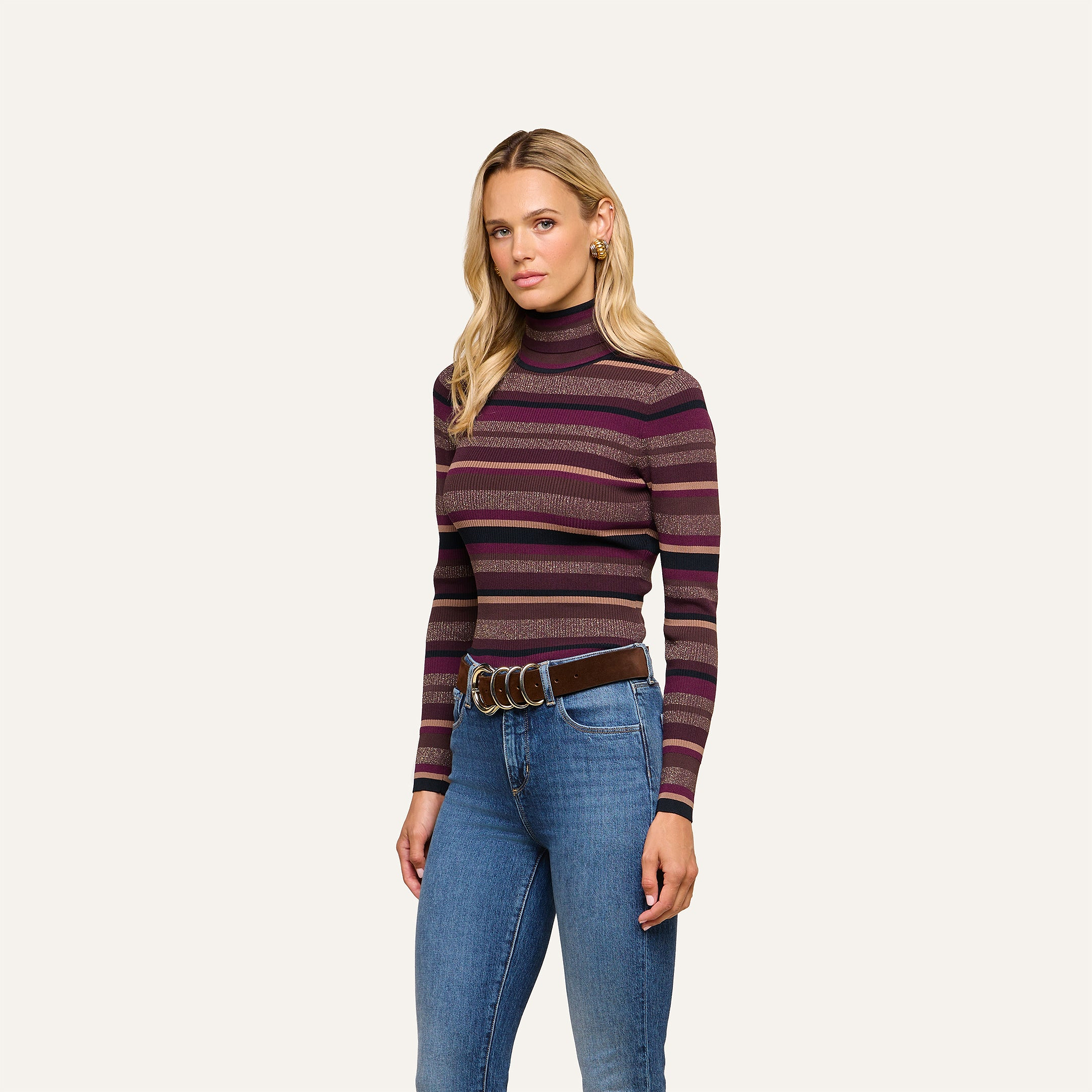 A woman with long blonde hair and a neutral expression stands against a plain background, modeling the Olene Striped Turtleneck—a ribbed knit sweater in shades of purple and pink—paired with blue jeans and a brown belt for an effortless fall fashion look.