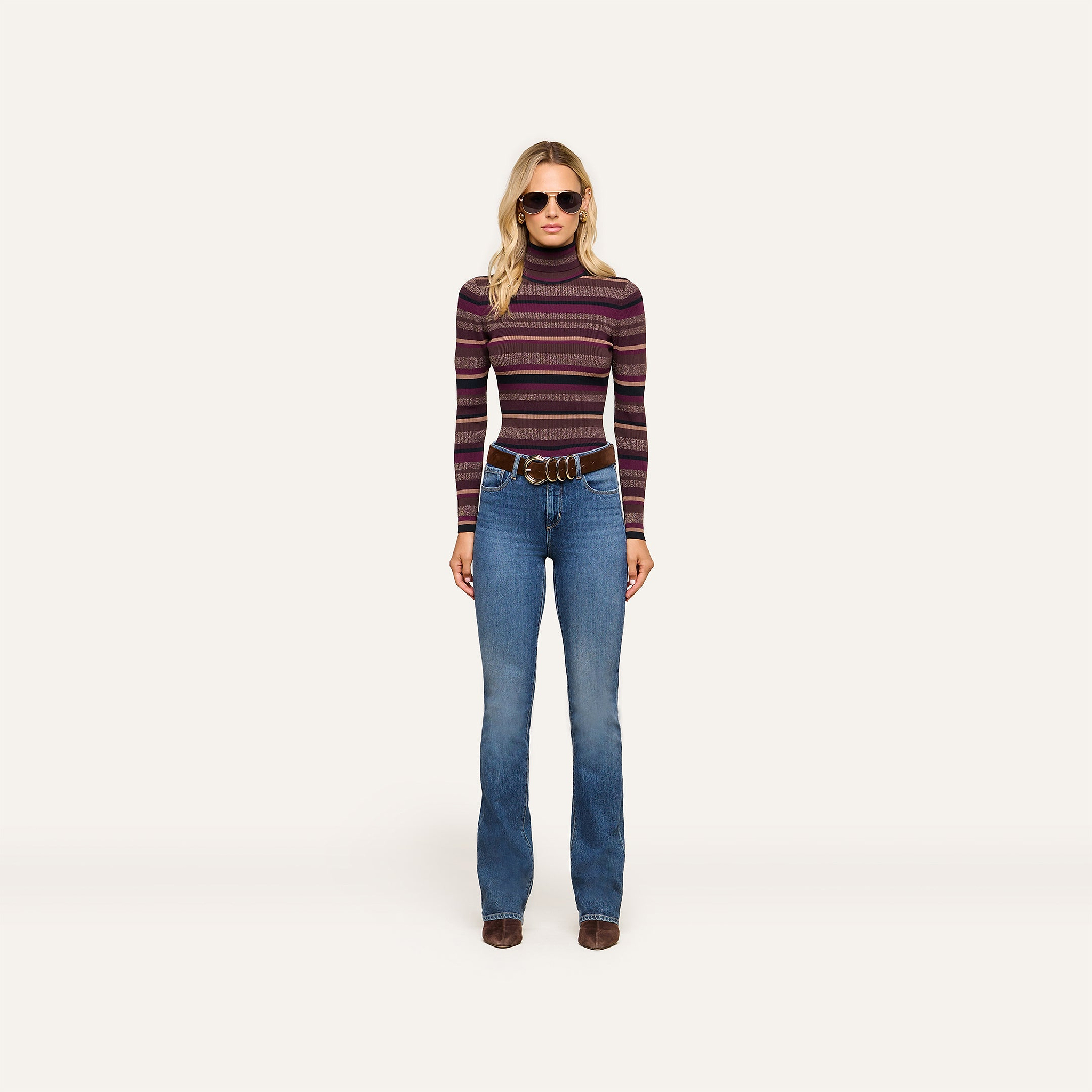 Wearing the Olene Striped Turtleneck with blue flared jeans, a wide brown belt, brown shoes, and stylish sunglasses, the woman stands against a plain light background—an effortless nod to classic fall fashion.