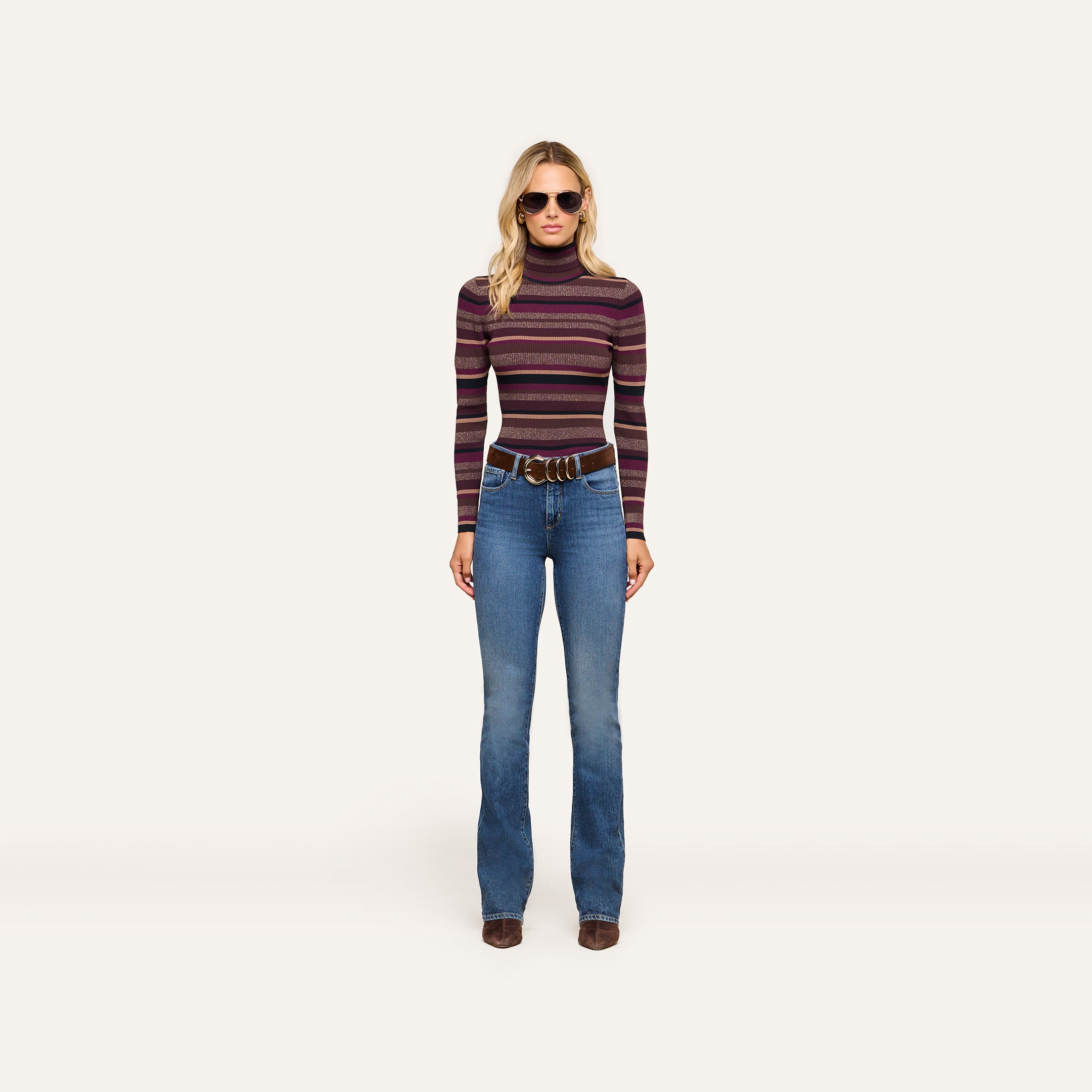 Wearing the Olene Striped Turtleneck with blue flared jeans, a wide brown belt, brown shoes, and stylish sunglasses, the woman stands against a plain light background—an effortless nod to classic fall fashion.