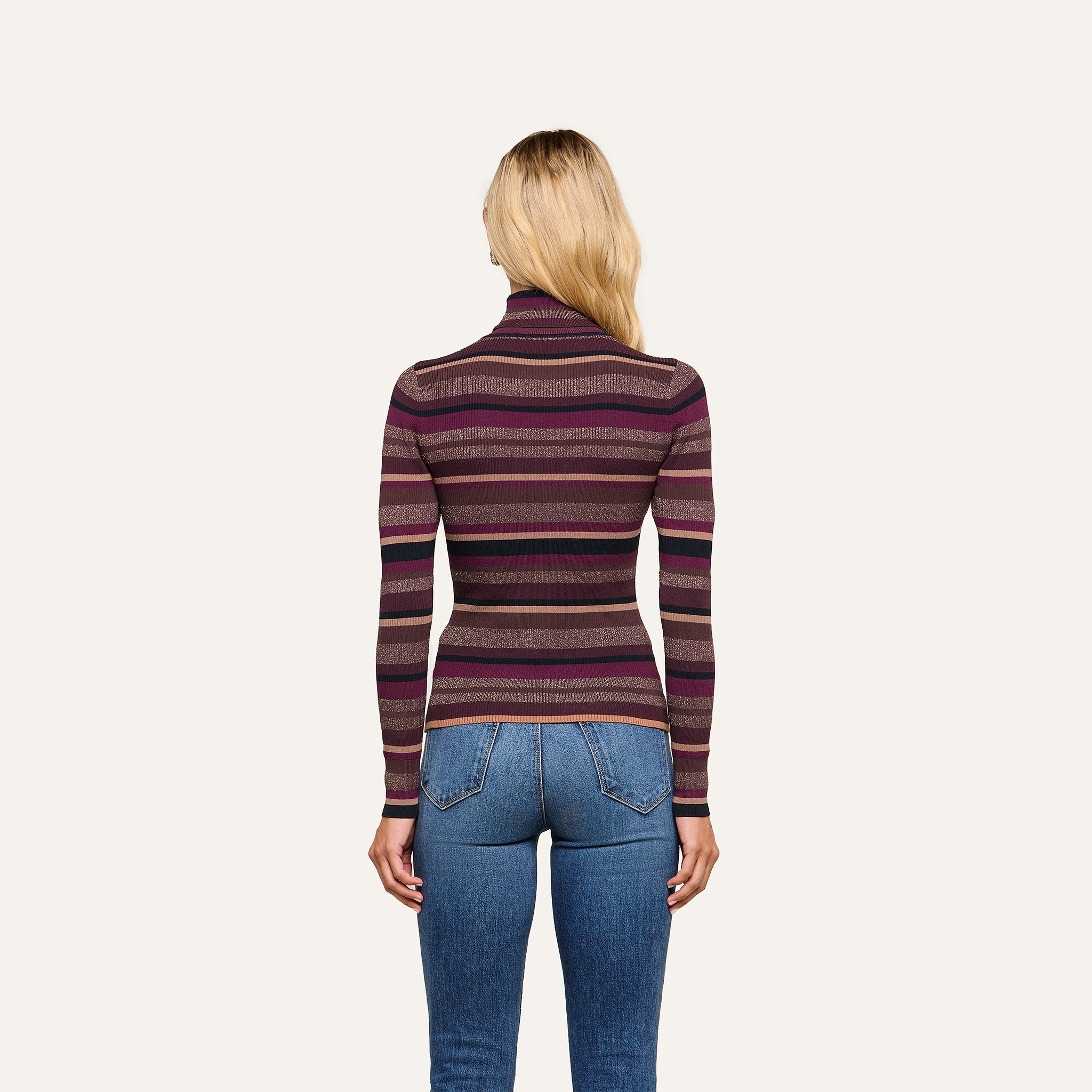A person with long blonde hair, wearing the Olene Striped Turtleneck—a ribbed knit sweater in shades of purple—paired with blue jeans, stands facing away from the camera against a plain background for an effortlessly stylish fall look.