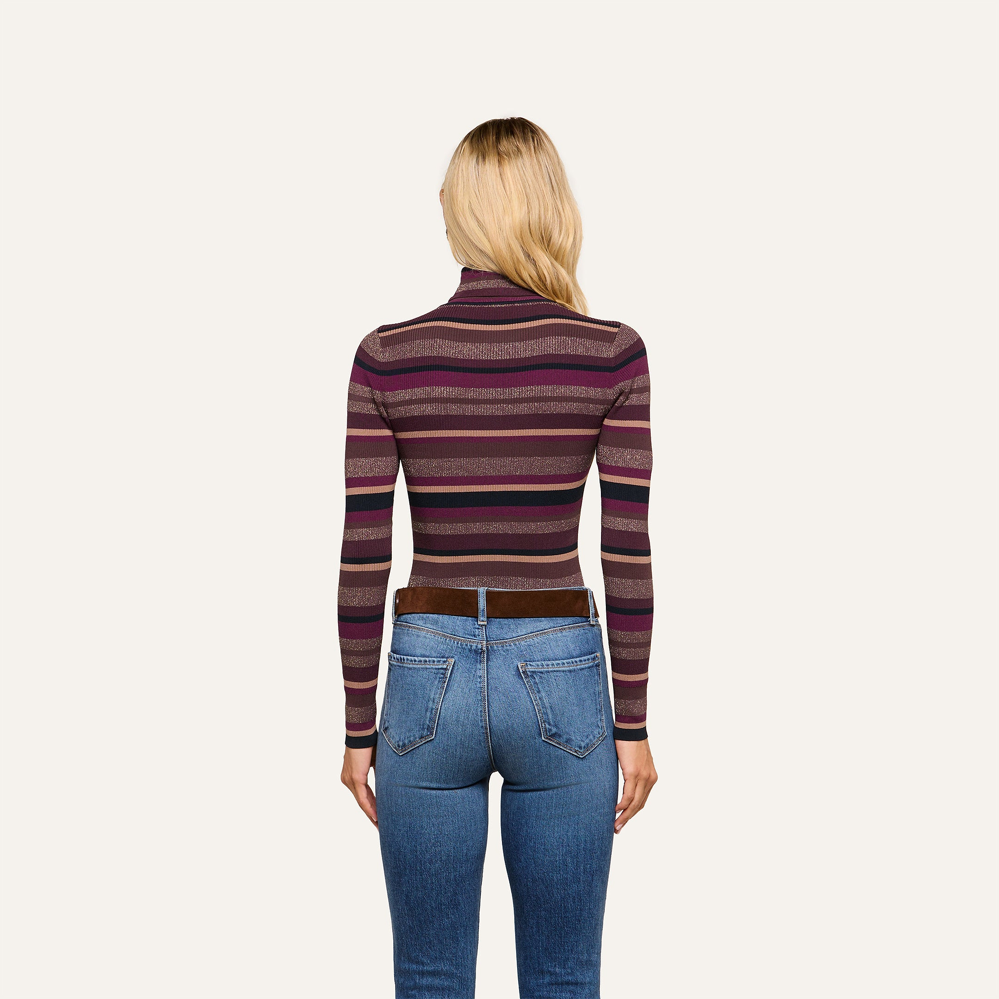 Wearing the Olene Striped Turtleneck in maroon and purple stripes paired with blue jeans, a person with long blonde hair stands facing away from the camera against a plain light background—an effortlessly stylish look ideal for fall fashion.