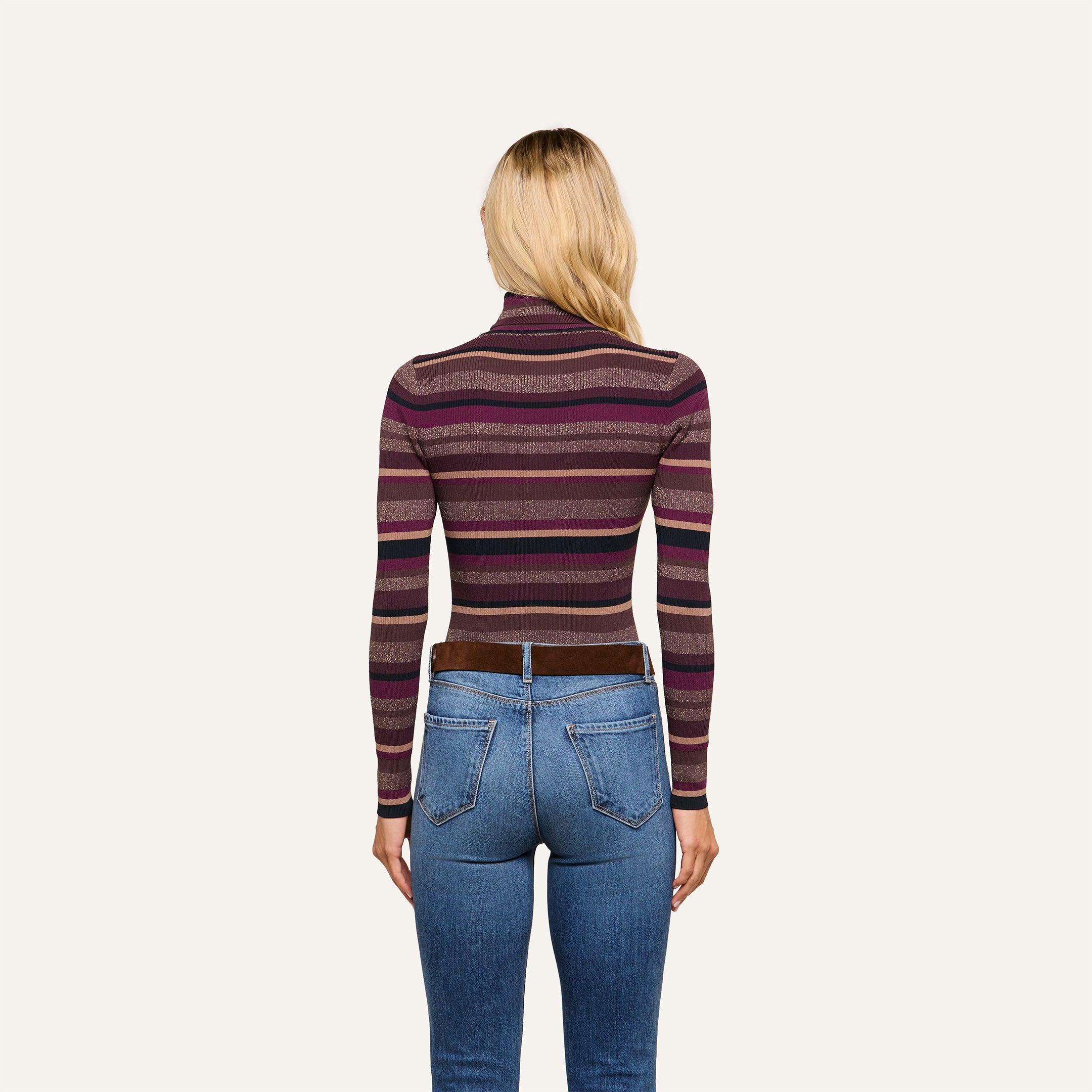 Wearing the Olene Striped Turtleneck in maroon and purple stripes paired with blue jeans, a person with long blonde hair stands facing away from the camera against a plain light background—an effortlessly stylish look ideal for fall fashion.