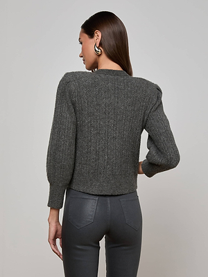 A woman with straight brown hair models the L'AGENCE Talulla Cardigan—featuring jeweled buttons—paired with high-waisted charcoal pants. She poses back to camera against a light gray background, highlighting the cardigan's textured knit design.