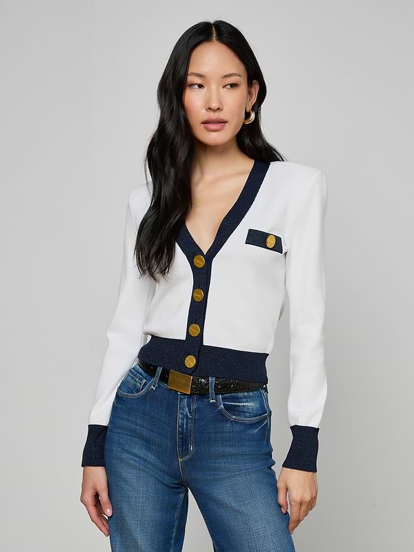 A woman with long black hair wears the Taya Cardigan, a chic Parisian piece with navy and metallic trim and gold buttons, styled with blue jeans and gold hoop earrings, set against a plain gray background.