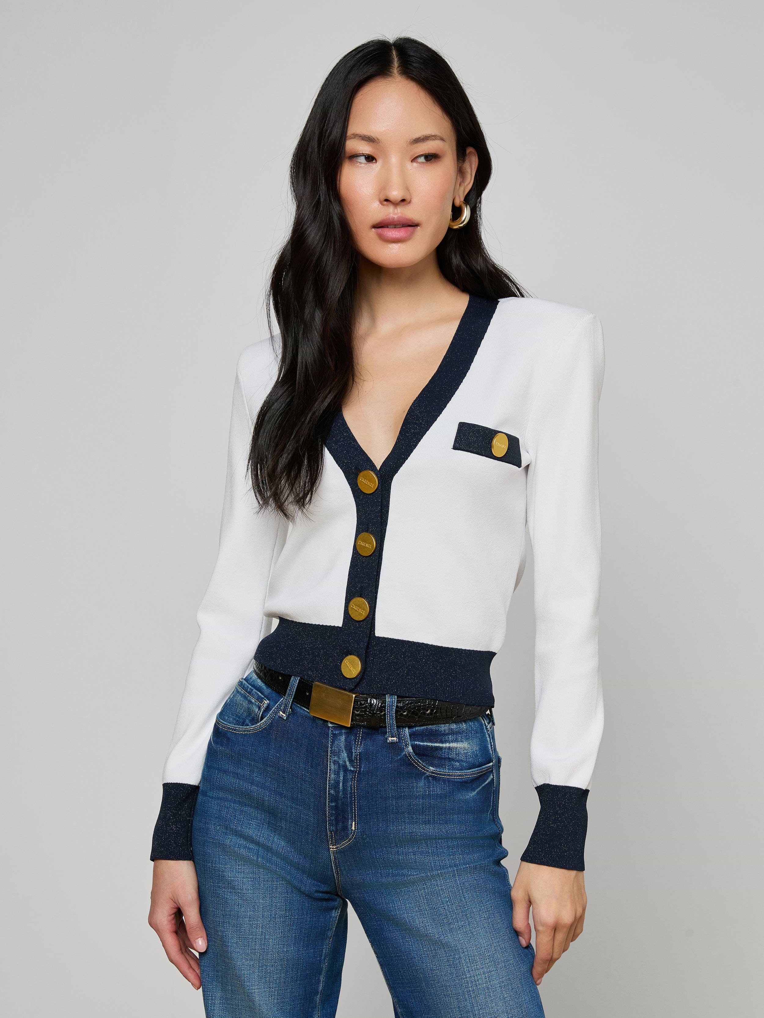 A woman with long black hair wears the Taya Cardigan, a chic Parisian piece with navy and metallic trim and gold buttons, styled with blue jeans and gold hoop earrings, set against a plain gray background.