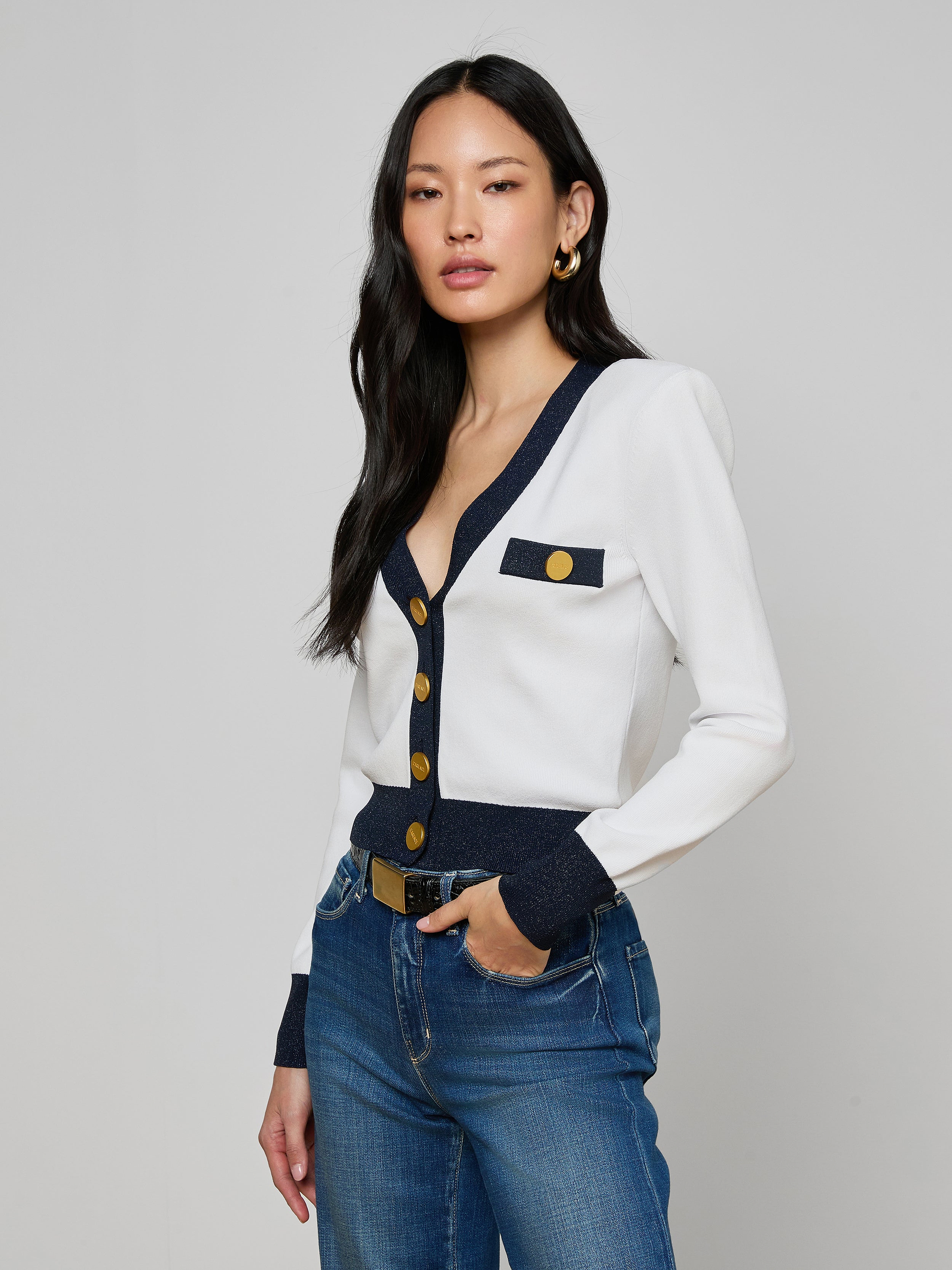 A woman with long black hair models the Taya Cardigan—Parisian-inspired in white with dark and metallic trim and gold buttons—paired with blue jeans and gold hoop earrings, standing before a plain light gray background.