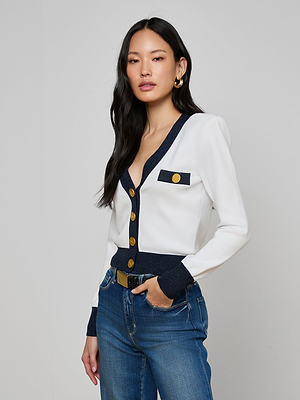 A woman with long black hair models the Taya Cardigan—Parisian-inspired in white with dark and metallic trim and gold buttons—paired with blue jeans and gold hoop earrings, standing before a plain light gray background.