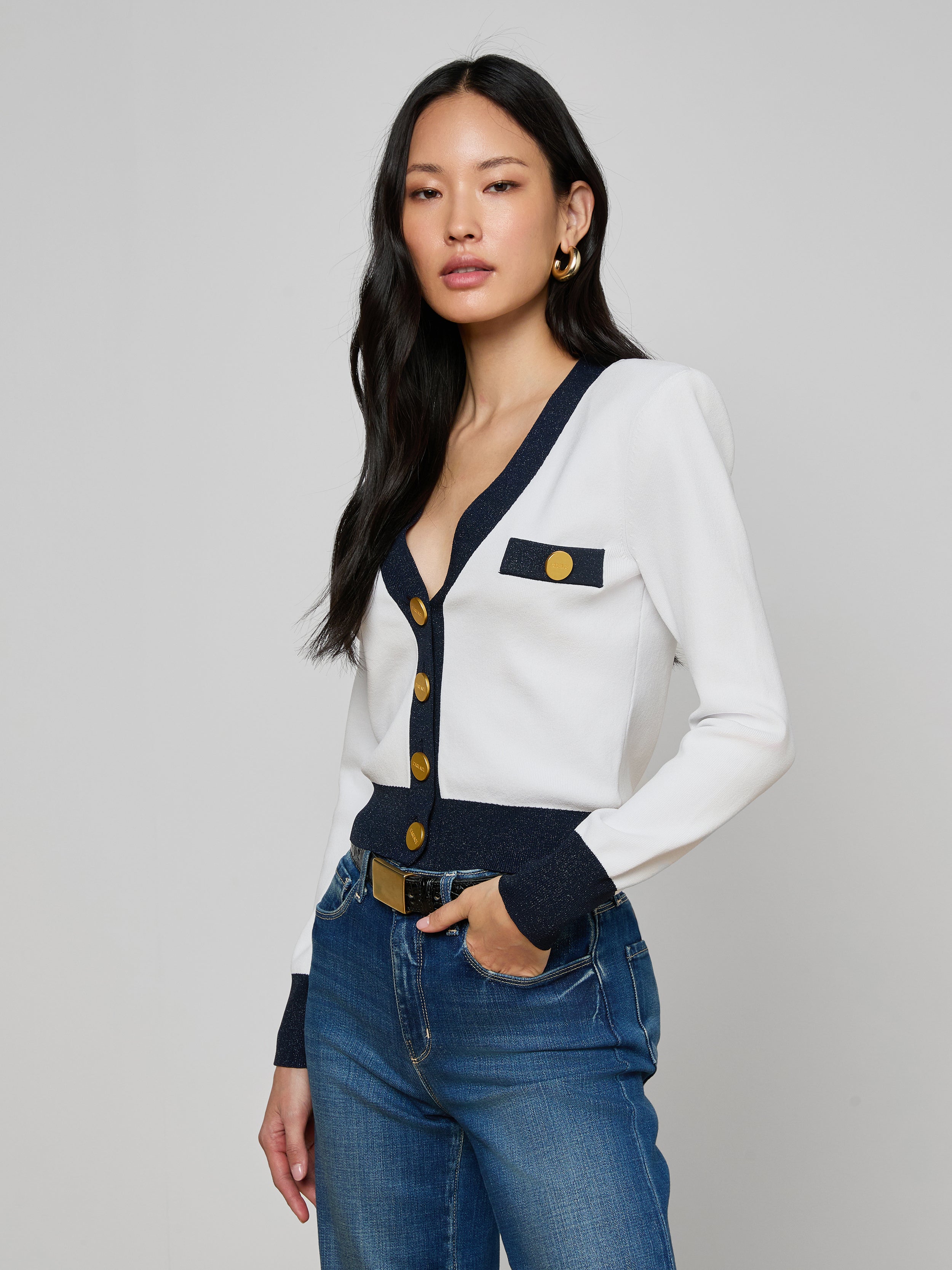 A woman with long black hair models the Taya Cardigan—Parisian-inspired in white with dark and metallic trim and gold buttons—paired with blue jeans and gold hoop earrings, standing before a plain light gray background.