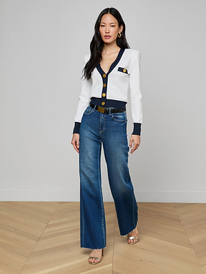 A woman with long dark hair poses indoors on a wooden floor, wearing the Taya Cardigan with black and metallic trim, gold buttons, high-waisted wide-leg blue jeans, and open-toed heels against a plain background.