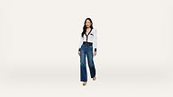 A woman stands against a plain white background, wearing a white blouse with black trim and buttons, blue wide-leg jeans, a black belt, and beige heels.