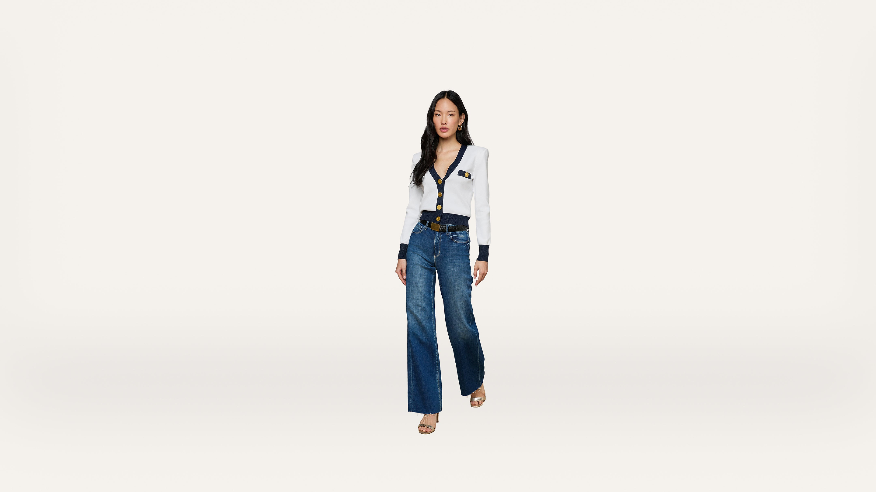 A woman stands against a plain white background, wearing a white blouse with black trim and buttons, blue wide-leg jeans, a black belt, and beige heels.
