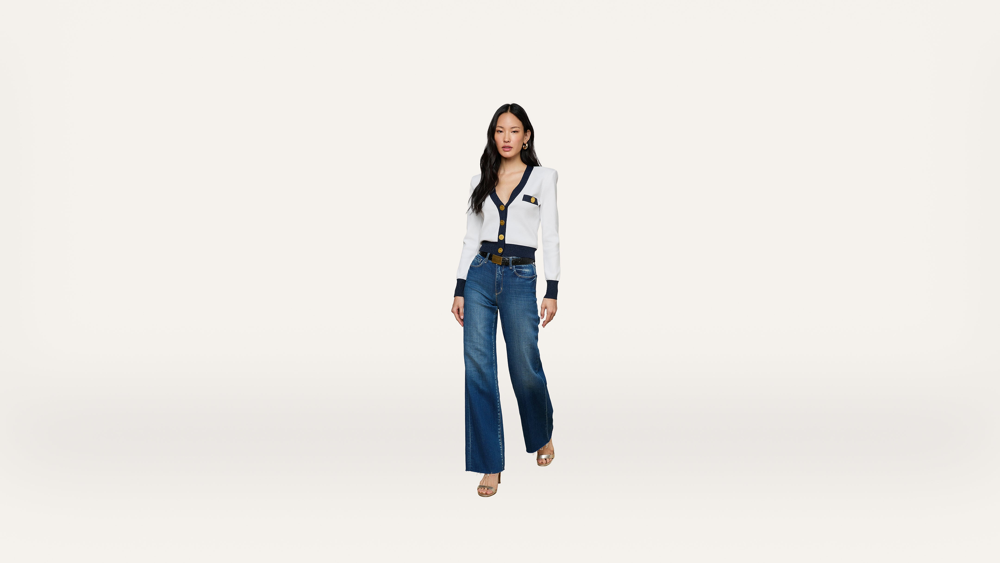 A woman stands against a plain white background, wearing a white blouse with black trim and buttons, blue wide-leg jeans, a black belt, and beige heels.