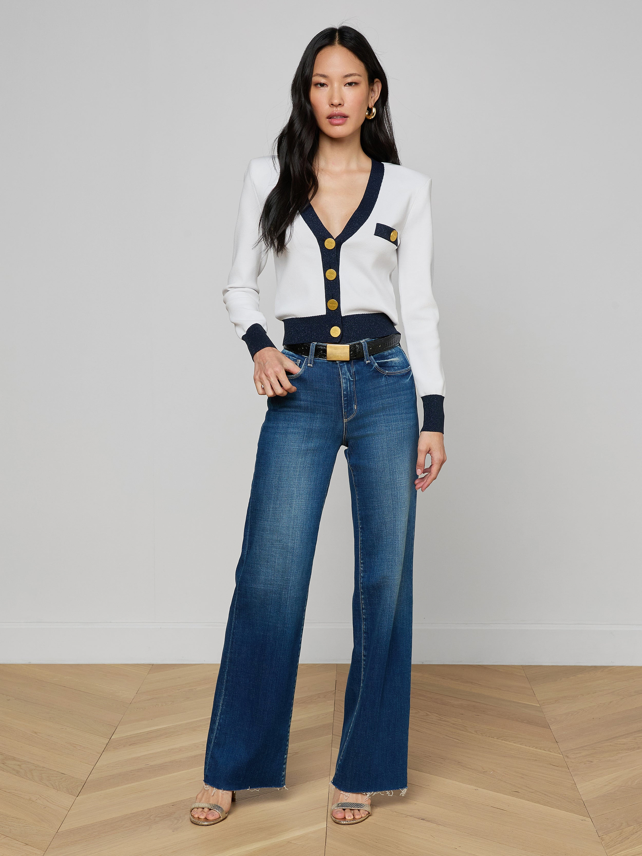 A woman stands indoors on a wood floor, wearing the Taya Cardigan with black and metallic trim and gold buttons, blue high-waisted wide-leg jeans, gold earrings, and open-toed heels. She poses facing forward with her hand by her side.