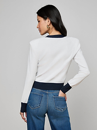 A woman with long dark hair, gold hoop earrings, and blue jeans models the Taya Cardigan—white with metallic trim and dark cuffs—standing with her back to the camera and left hand in her pocket against a plain gray background.