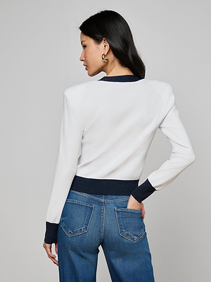 A woman with long dark hair, gold hoop earrings, and blue jeans models the Taya Cardigan—white with metallic trim and dark cuffs—standing with her back to the camera and left hand in her pocket against a plain gray background.