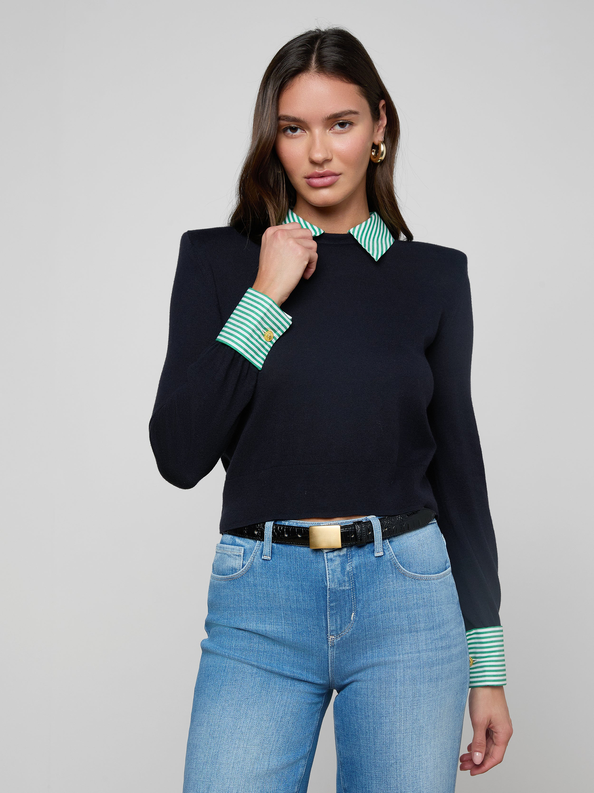 A woman stands against a plain background wearing the April Poplin Collar Pullover, featuring a navy featherweight knit with green and white striped collar and cuffs, paired with light blue jeans, a black belt with gold buckle, and gold hoop earrings.