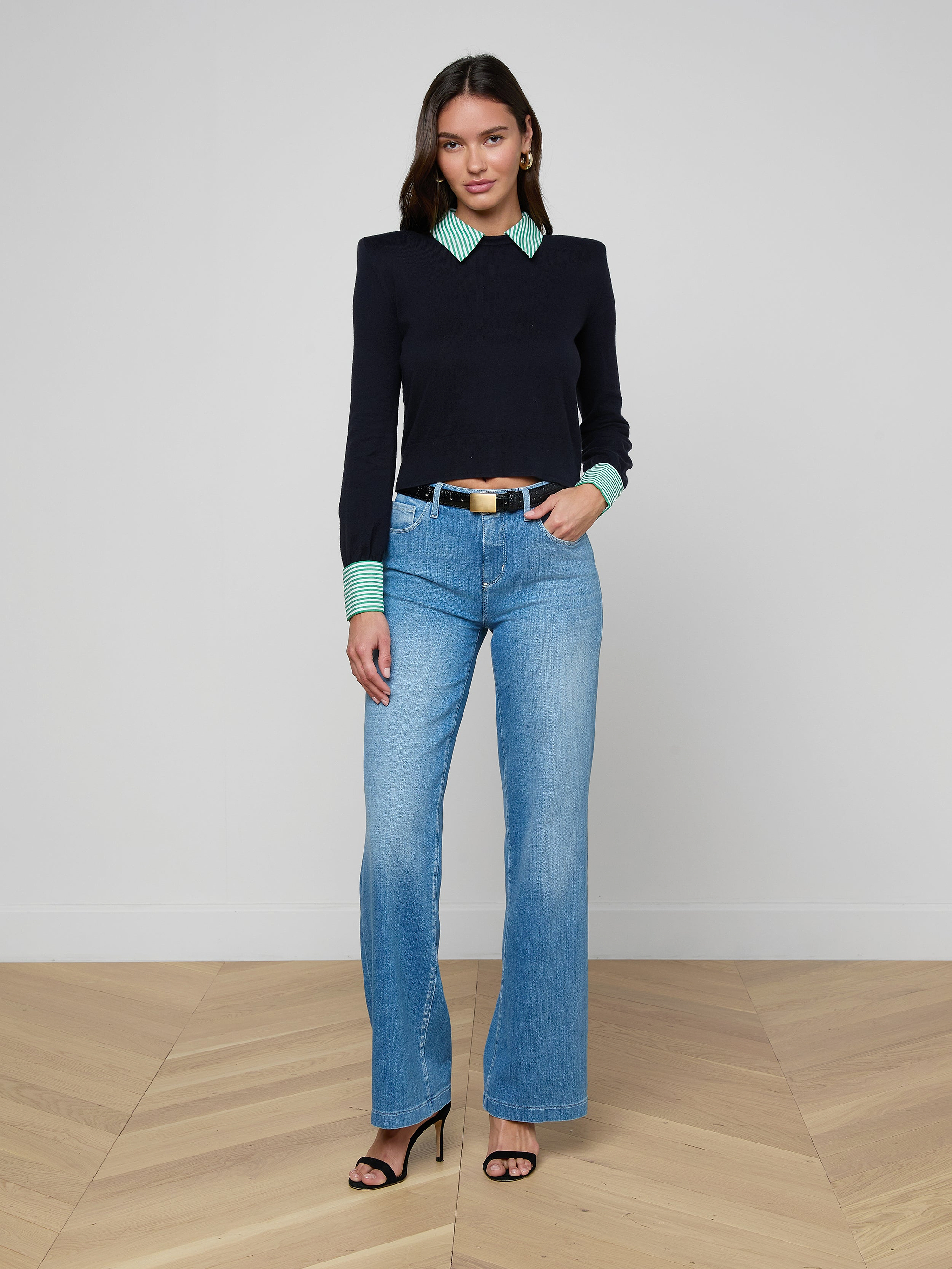 A woman with long hair stands indoors on a wooden floor, wearing the April Poplin Collar Pullover, light blue jeans, and black high-heeled sandals. She faces the camera with a slight smile.