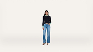 A woman stands facing forward, wearing a black sweater over a green shirt, blue jeans, and black heeled sandals, against a plain white background.
